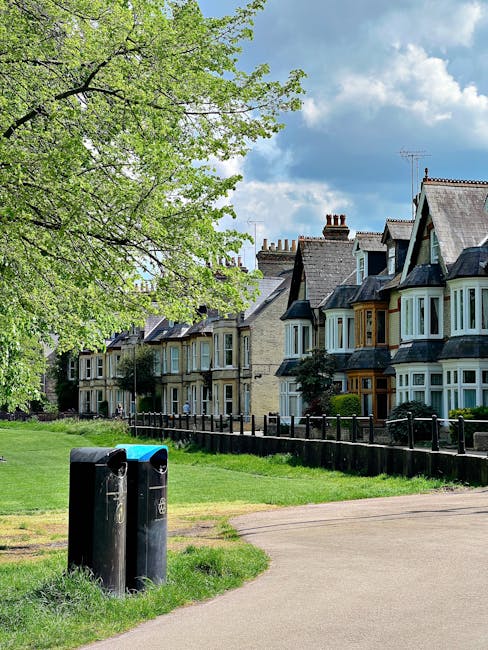 The image depicts a suburban street scene featuring a row of traditional terraced houses with varied architectural details, including bay windows and multiple chimneys, situated behind a well-maintained wooden fence. In the foreground, two black rubbish bins with slightly reflective surfaces stand on a grassy verge near a paved pathway, which curves gently to the right. To the left, a large leafy tree with fresh, light green foliage extends its branches over the scene, contrasting with the partly cloudy sky overhead that has patches of blue and white clouds. The environment suggests a peaceful residential area, where private waste collection or on-site clearance services, such as those offered by Rubbish Collection Chiswick, could be relevant for managing household rubbish. The overall atmosphere is bright and clear, emphasizing the tidy appearance of the neighborhood and the potential suitability for independent waste management solutions.