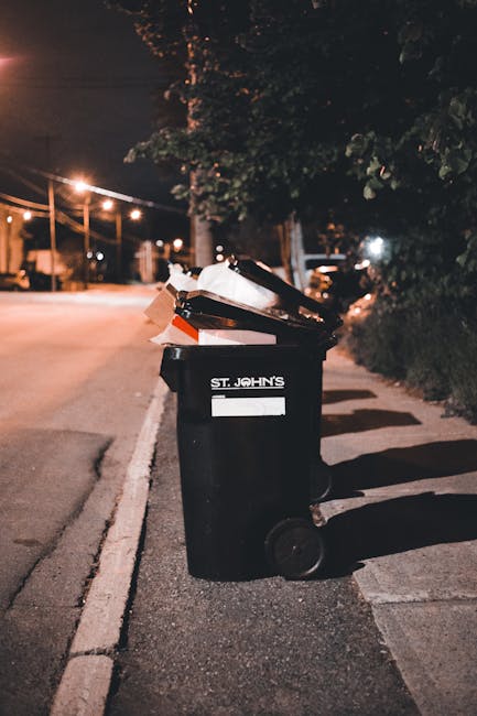 A black wheeled rubbish bin with a white label and the words 'St. John's' printed on the front, positioned on a sidewalk beside a curb in an urban residential area at night. The bin's lid is open, revealing a mix of waste materials including cardboard boxes, paper, and plastic wrappers. Surrounding the bin are shadows cast by nearby streetlights, with trees and houses visible in the background, illuminated by a warm, amber glow. The pavement and street appear wet, reflecting the streetlights, indicating recent rain. The scene illustrates typical private waste disposal practices in a suburban setting, with the bin ready for municipal collection or alternative waste handling services. Rubbish Collection Chiswick often provides similar on-site clearance or independent rubbish removal options for residents. The overall composition emphasizes the importance of proper waste management in urban environments, especially when opting for private rubbish collection services instead of council-provided refuse collection.