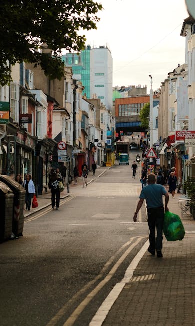 A bustling urban street scene shown with a variety of buildings lining both sides, including traditional terraced shops with small frontages, some with awnings and signage. Pedestrians are walking along the pavement, some carrying shopping bags or pushing strollers, while others are crossing the street at a designated pedestrian crossing. In the foreground, a man dressed in a turquoise shirt is walking away from the camera, holding a bright green wheeled bag, possibly containing waste or recyclables, which suggests activity related to rubbish removal or private waste collection. The street surface is a mix of paving and road markings, with a double white line at the edge indicating a no-parking zone. The background features a mix of older buildings and taller modern structures, with some visible signage and shop fronts, hinting at a commercial district with active foot traffic. Overhanging tree branches partially shade the scene, and the overall atmosphere suggests a typical day in a busy town center, where independent waste handling services like Rubbish Collection Chiswick might operate to support local rubbish removal needs in such environments.
