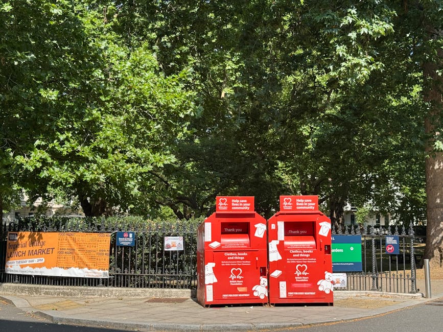 Two red collection boxes positioned on a paved sidewalk in front of a black metal fence, which encloses a park area with dense green trees and foliage in the background. The boxes are designated for clothing, books, and general items, featuring white icons of a T-shirt, book, and box, along with white text indicating their purpose. Each box has a small top panel with a white heart logo and a message about helping the community. The scene is outdoors, illuminated by natural daylight, with the boxes standing upright and closely positioned side by side on the corner of a street, suggesting a location for private or independent waste handling as part of community collection efforts. An orange vertical banner advertising a local lunch market is attached to the fence on the left, partially visible behind the fence, with some trees towering above, creating a shaded environment typical of urban parks or community areas. Rubbish Collection Chiswick occasionally handles civic or charity collections of these sorts, facilitating environmentally responsible disposal outside of official local authority services.