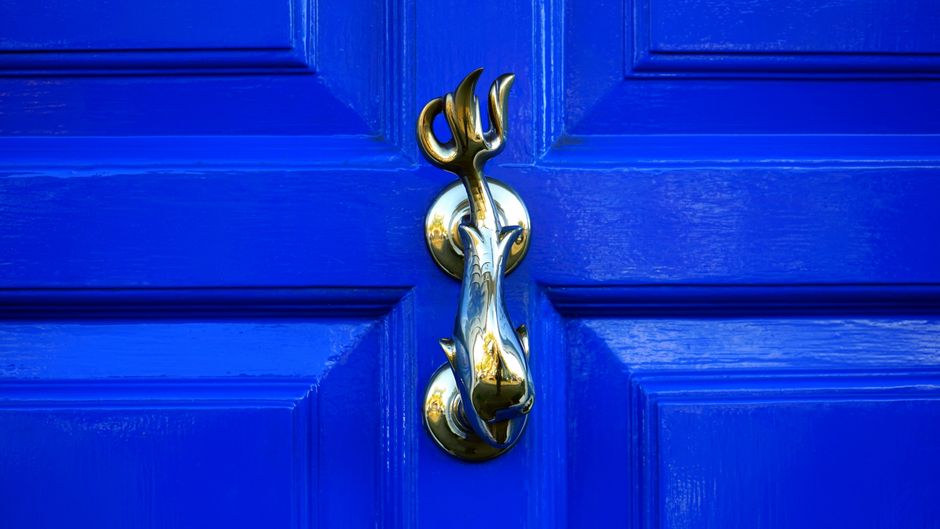A close-up view of a shiny, gold-toned door knocker in the shape of a dog’s head with a small, three-pronged trident above it, mounted on a vibrant blue-painted wooden door. The door features rectangular paneling around the knocker, and the surface has a glossy finish that reflects light subtly. The door appears to be part of an exterior structure, possibly near a garden or pathway, with a clean and well-maintained appearance. This scene illustrates a decorative door accessory typically associated with private properties, providing a visual example of alternative access points for rubbish collection or property entry in the context of local waste disposal services. Rubbish Collection Chiswick occasionally services properties with distinctive exterior fixtures like this, supporting private waste handling solutions rather than relying solely on municipal rubbish collection networks.