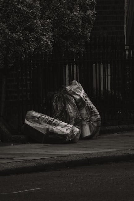 Two large black plastic rubbish bags, partially torn and crumpled, rest against a black metal fence on a paved pavement in an outdoor urban environment. The bags appear to contain household waste and are situated near a sidewalk edge. Behind the fence, there are dense green foliage and trees, indicating a residential or garden boundary, with some building structures partially visible in the background. The scene is illuminated by natural daylight, creating subtle shadows and highlights on the plastic surface, emphasizing the textures and creases of the bags. The image relates to waste management and private rubbish collection, highlighting the type of waste disposal often handled through independent rubbish removal services such as those offered by Rubbish Collection Chiswick, emphasizing the need for on-site clearance and timely rubbish removal solutions.