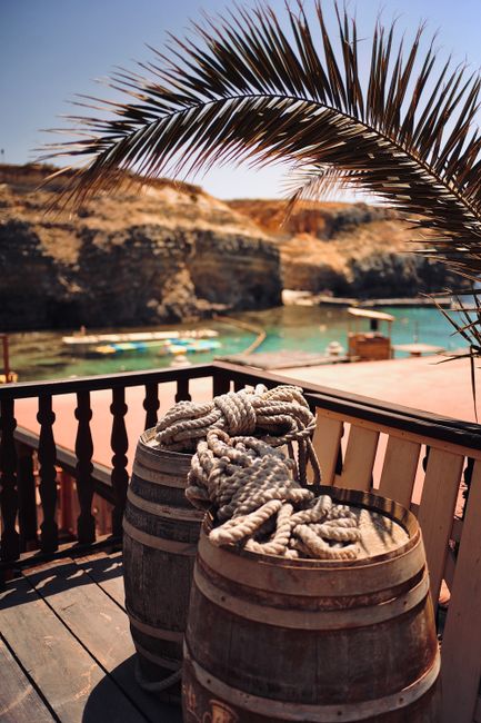 In the foreground, two wooden barrels are positioned on a wooden deck or balcony, each topped with thick, white, coiled ropes that appear to be made of natural fiber, possibly for maritime purposes. The barrels display weathered textures, with visible wood grain and metal bands, evoking a rustic beachside or seaside theme. Behind the barrels, a wooden railing with vertical balusters runs along the edge of the deck, partially obscuring a view of a calm, turquoise body of water with several small boats floating near the shoreline. In the background, sandy cliffs rise along the coast, featuring patches of sparse vegetation, and the sky above is clear and blue, suggesting a sunny day. A large, arching palm frond extends across the top portion of the image, its spiky, dark green leaves contrasting against the sky and providing a tropical atmosphere. The overall scene suggests an outdoor location close to the water, possibly a seaside cafe or coastal retreat, with natural elements and casual maritime accessories emphasizing a relaxed, beachside setting. This scene aligns with the context of waste management or rubbish removal services for leisure or hospitality environments, as offered by Rubbish Collection Chiswick, though no waste materials are visible in the image.