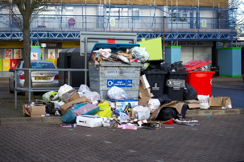 A large pile of mixed rubbish and waste materials accumulated in a city centre setting, positioned on a paved sidewalk alongside a metal railing. The refuse includes overflowing black and black-and-white bin bags, open cardboard boxes, crumpled paper, plastic containers, and discarded packaging materials. A prominent, blue-lid mixed waste bin for paper and cardboard is partially open, revealing some paper waste spilling out, with its surface marked by labels. To the right, a bright red wheelie bin, also overflowing, contains visible plastic and paper debris. Nearby, other black bins and various loose rubbish are scattered around, some resting directly on the ground, with packaging and waste extended onto the pavement. The background features a commercial building with blue scaffolding and protective netting, alongside storefronts with partially visible signage, and a parked car is located behind the rubbish pile. The scene is illuminated by daylight, emphasizing the disorder and typical of private waste collection areas, with Rubbish Collection Chiswick often providing alternative rubbish removal services for such excess or non-regular waste disposal scenarios.