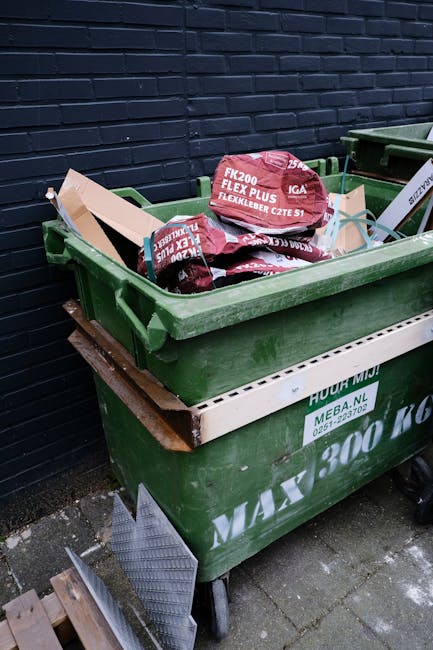 A large green wheeled skip filled with red and black bags of construction or garden waste, placed on a paved surface beside a dark brick wall. The bags are labeled with brand names and product details. The skip is positioned next to another similar container with partially visible contents and a white barrier or platform attached to the front. The environment appears to be an outdoor area, likely adjacent to a property or waste management site, with the scene suggesting private waste handling or on-site clearance services offered by Rubbish Collection Chiswick. The skip's durable plastic construction has a textured surface, and the bags inside are made of flexible material, with some stacked and leaning slightly against each other. The lighting indicates natural daylight, emphasizing the contrast between the dark wall, green skip, and the red bags. This image visually supports independent rubbish collection and waste disposal services within the context of alternative waste handling solutions in the W4 Chiswick area.