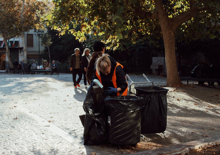 A black wheelie bin labeled 'St. John’s' with a white rectangular space beneath the label, positioned on a pavement during nighttime. The bin is filled with discarded flat cardboard boxes, paper, and plastic packaging, which are slightly overflowing and protruding from the open lid. The scene is illuminated by nearby streetlights, creating warm light that highlights the texture of the bin’s surface and the surrounding asphalt. In the background, blurred streetlights and faint outlines of trees and parked cars can be seen, indicating a residential area. The pavement edge runs parallel to the bin, with some shadows cast by the tree leaves and the bin itself. This scene subtly reflects the concept of private or alternative waste handling, with the bin capacity utilized for refuse collection typical of rubbish removal services like Rubbish Collection Chiswick, especially in a neighborhood setting fitting for local rubbish collection and disposal efforts.